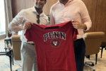 Two people smiling and holding a red T-shirt with "PENN" and a crest design, representing the University of Pennsylvania, in an office setting.